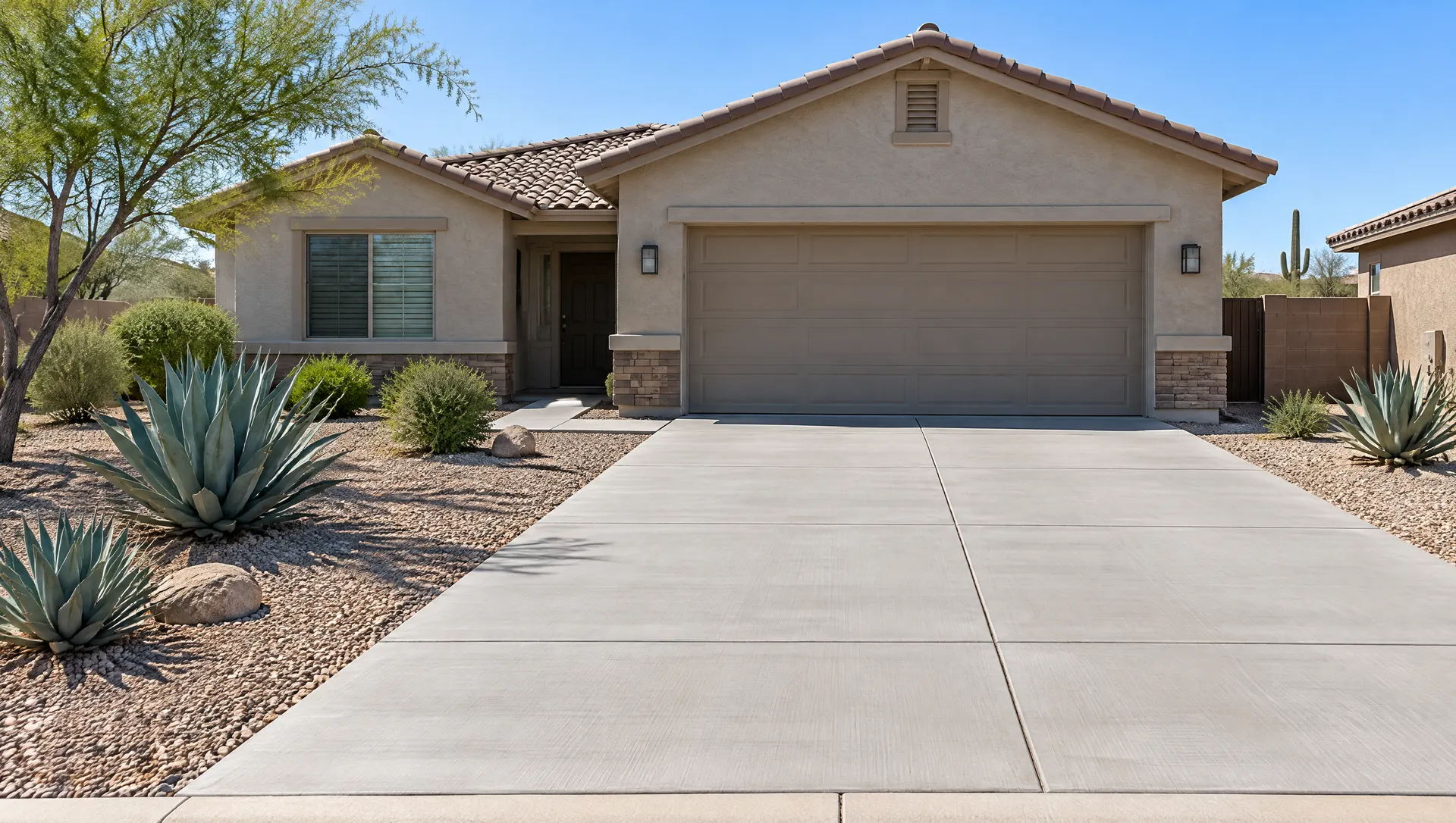 Brand new wide concrete driveway with a smooth broom finish and straight control joints in front of a Phoenix ranch home with a two-car garage, gravel front yard and agave plants, bright clear morning